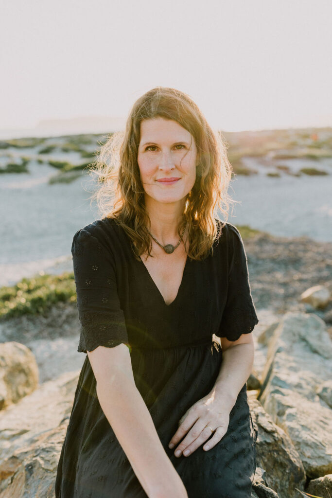 Lauren sits smiling with her back to the ocean. Her hair is dark blonde and wavy, and it falls just below her shoulders. She's wearing a navy blue dress with a v-neck and scalloped sleeves that almost reach her elbow. She's wearing a bronze and silver necklace.